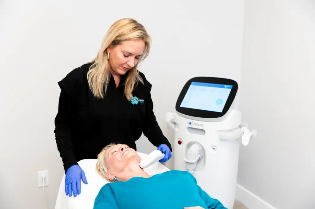 A female Restoration MedSpa provider, wearing black scrubs and blue gloves, gently presses the Sofwave device to a female client's neck for skin tightening in Greensboro.
