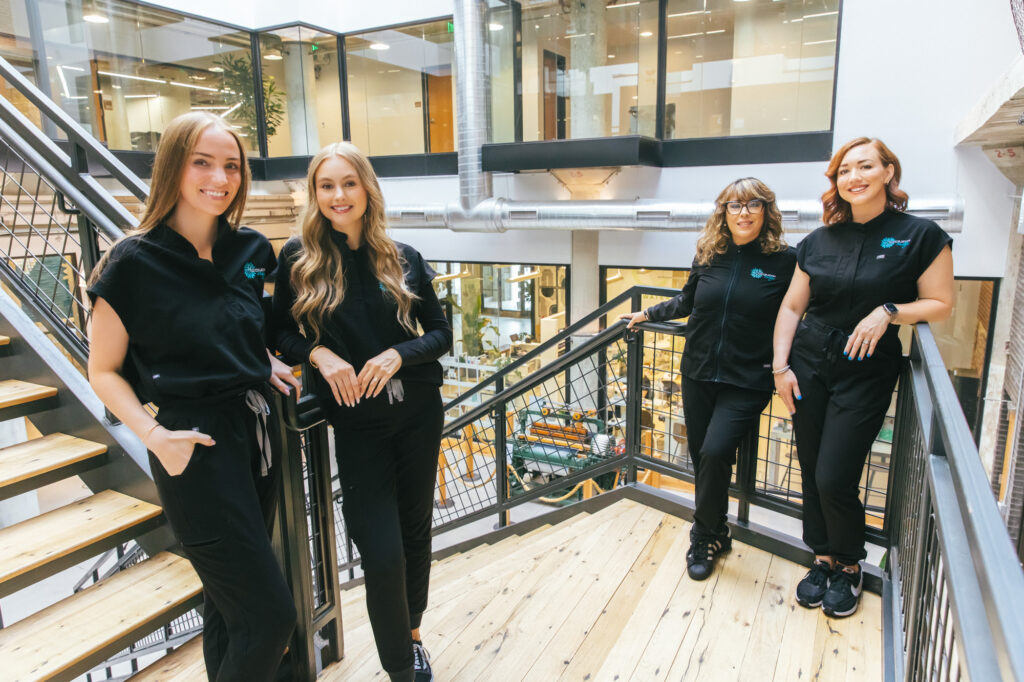 4 female Restoration MedSpa providers stand on stair landing. They're each wearing black scrubs and smiling, ready to welcome clients to the best med spa in Greensboro.