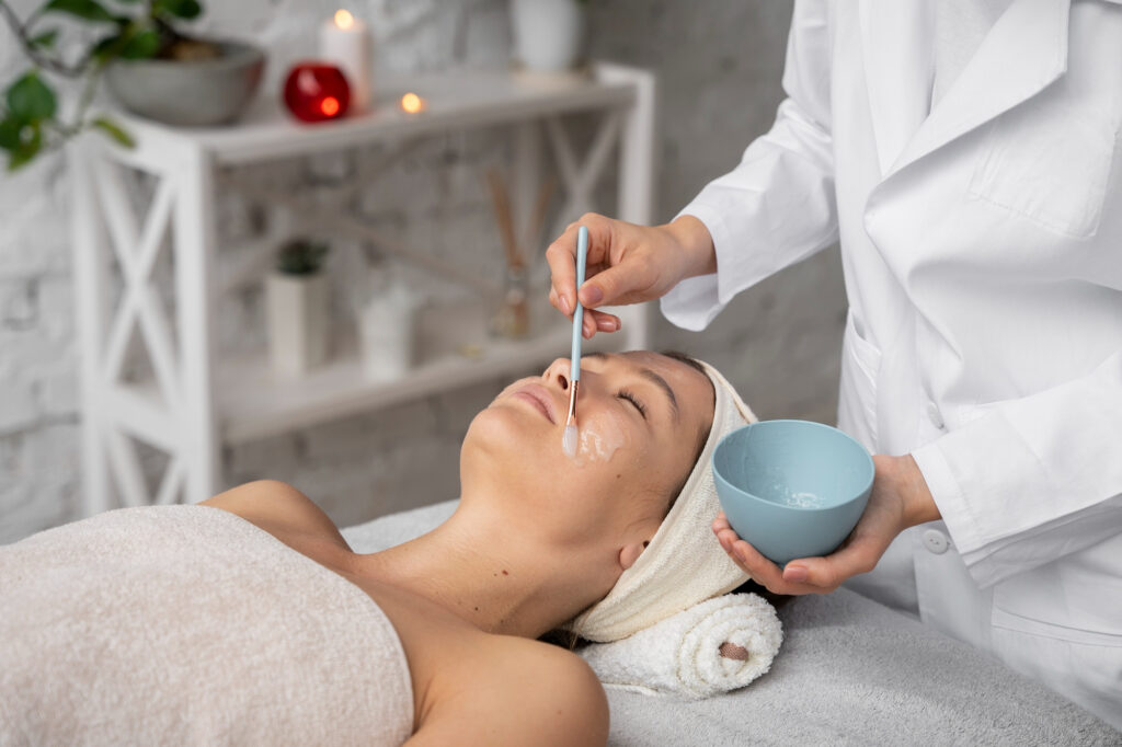  A woman lies on a treatment bed, covered in a grey towel, as a medical provider in a white coat applies an acid peel in Greensboro. There is a shelf with lit candles in the background. Acid peels remove layers of dead skin to reveal the new, brighter skin underneath.