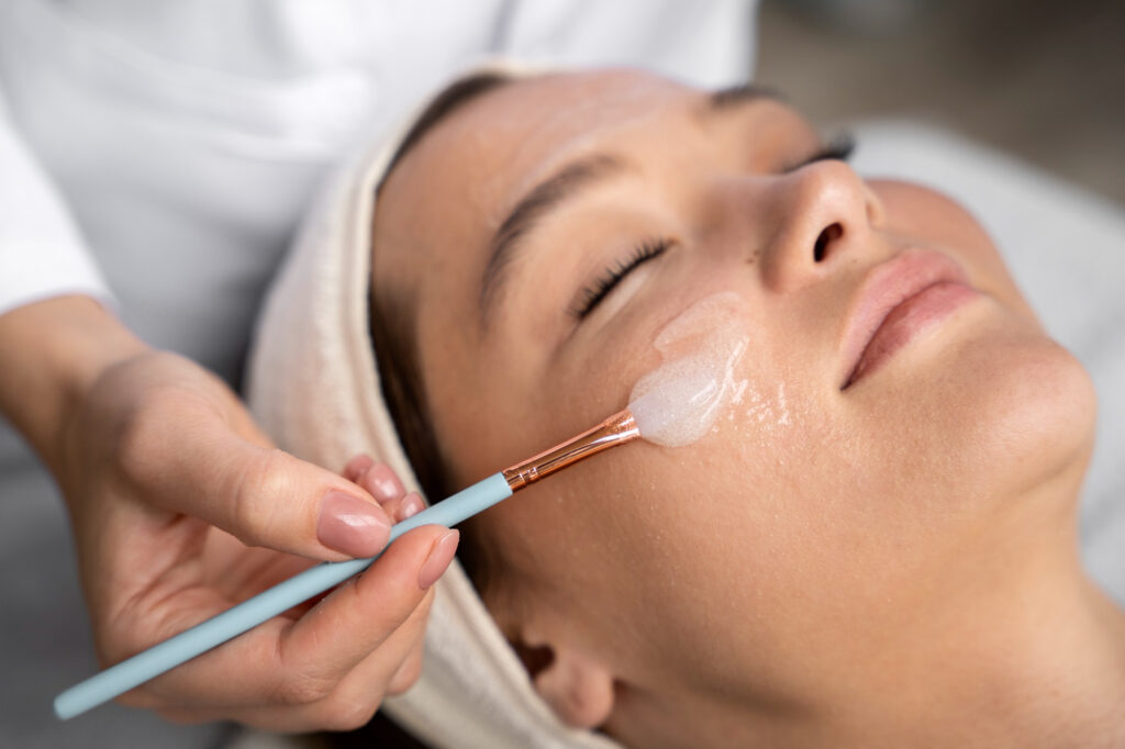A close-up on a female spa client's face. A medical provider, wearing a white coat, stands behind her, applying an acid peel in Greensboro. This peel, also known as a chemical peel, comes in three strengths: light, medium, and deep.