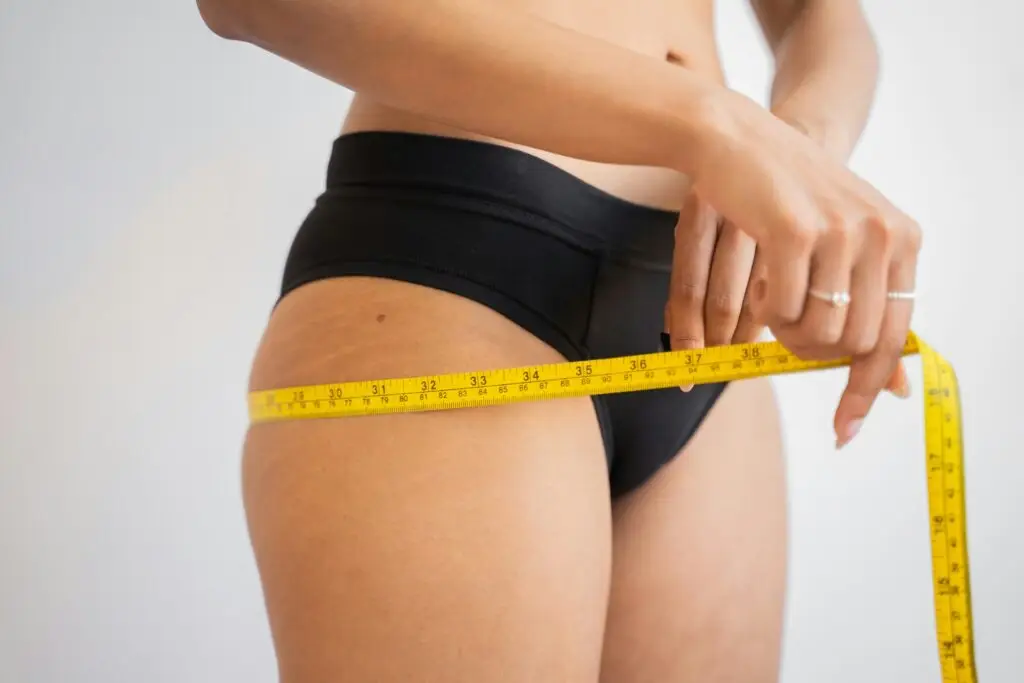 A female Restoration MedSpa client, wearing black briefs, measures her hips with a yellow cloth tape measure after receiving medically-guided weight loss injections. This option for body contouring in Winston-Salem can manage blood sugar, slow digestion, and alter hunger cues for sustainable weight loss.
